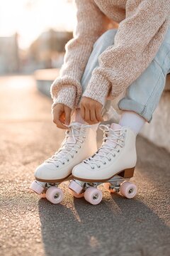 A girl sits on a bench in a park during the afternoon. She is tying her pink roller skates and wears a sweater. There are trees and sunlight in the background