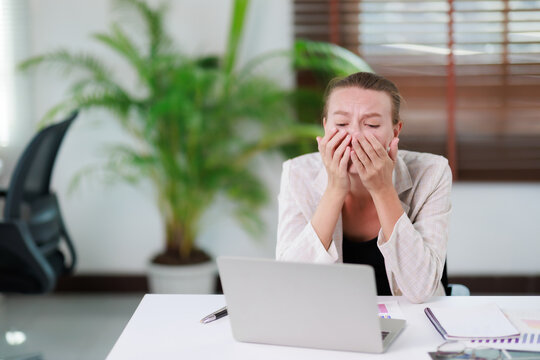 Stressed businesswoman feeling overwhelmed and frustrated, covering her face with hands while experiencing burnout and exhaustion at the office desk with a laptop