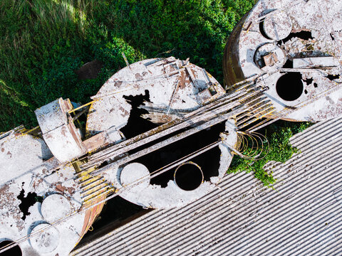 Aerial view of abandoned and decaying industrial grain silos with rusted metal surfaces and damaged corrugated roofs surrounded by overgrown vegetation in Latvia.