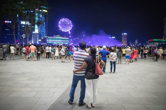 Couple stands embracing among crowd watching illuminated night light show with glowing sphere lantern and city skyline reflected in purple haze at Singapore waterfront promenade