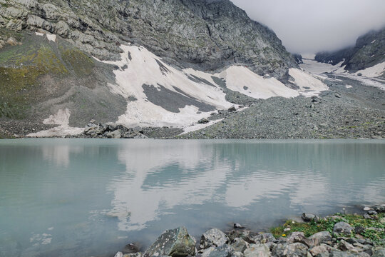 Milky waters of White Abudelauri Lake mirror lingering snow and misty cliffs below the Chaukhi massif in Georgia, creating a cold, peaceful and otherworldly mountain atmosphere