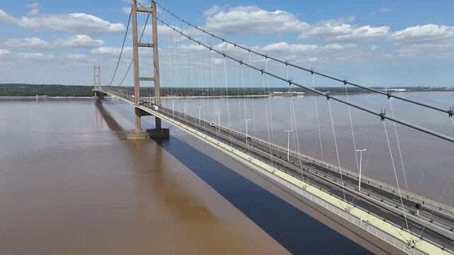 Bridge spans wide, muddy river under bright sky. Clouds drift above, adding soft, natural rhythm. Vehicles move steadily, connecting distant lands. Sky, water, and steel create calm, powerful scene