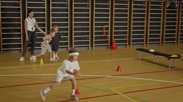 Caucasian children running relay race around cones in school gym while female coach supervising. Footage is ideal for promoting youth sports programs, physical education, and active lifestyles