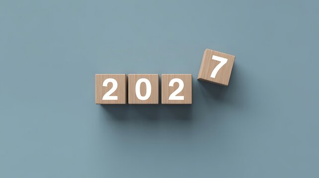 Wooden blocks displaying the year 2027 with the final number seven being placed into position on a plain grey background, representing future planning