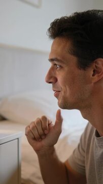 Vertical video: Speaking man in grey tee replying to device prompt at bed edge, dresser, copy space