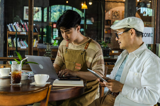 Modern small business digital management young entrepreneur using laptop and tablet planning, finance workflow efficiency technology cafe business operation, learning from senior cafe owner experience