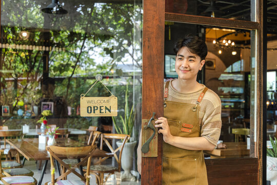 Young barista cafe owner smiling at entrance with open sign visible, confident entrepreneur representing hospitality service and customer readiness, positive mood in small business operation.