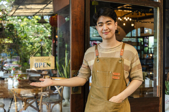 Young small business cafe owner barista open sign welcome gesture standing at entrance inviting customers into coffee shop friendly service concept and positive business presentation, cheerful worker