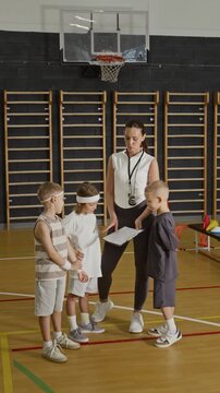 Vertical shot of young adult Caucasian female coach explaining sports strategy to three Caucasian kids in school gym. Footage is perfect for youth athletics and physical education promotions