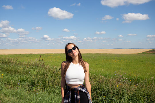smiling young woman in sunglasses standing in a green field under a blue summer sky, representing rural lifestyle, outdoor wellbeing, natural beauty and carefree freedom