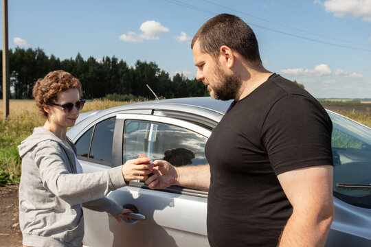 man accepts a car key from a woman beside a parked sedan in the countryside, suggesting vehicle rental, ownership transfer, transport agreement and secure personal mobility