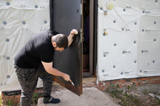 man scraping loose rust and deteriorated coating from an old entrance door during diy home renovation, preparing the steel surface for repainting and protection