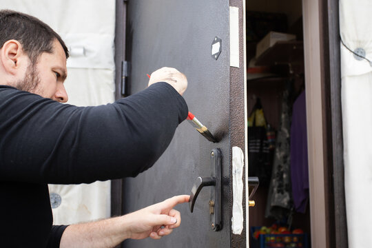 man carefully painting around the handle of an old steel entrance door, protecting masked hardware while restoring the exterior surface during diy home renovation