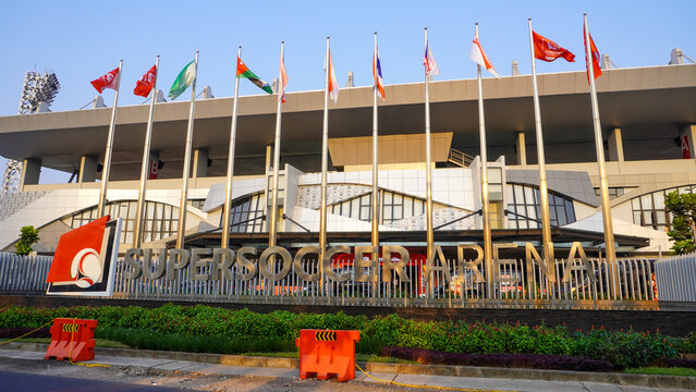 Exterior facade view of modern Super Soccer Arena featuring large metallic entrance signage, rows of tall flagpoles with flags waving, decorative gardens, and orange road barriers on sunny day.