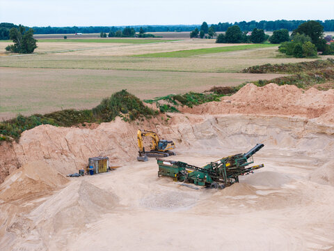 Aerial view of a gravel pit with an excavator and a mobile screening machine surrounded by fields in Slampe parish, Tukums Municipality, Latvia.