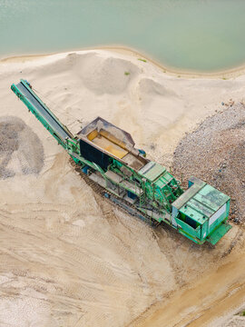 Aerial view of a green industrial gravel crushing machine operating in a sandy quarry near a water pond in Slampe parish, Tukums Municipality, Latvia.