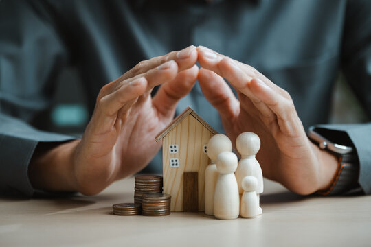 Home and family figures are sheltered by careful hands, visualizing property insurance, mortgage responsibility, and protected investment. Family financial, planning; family focus.