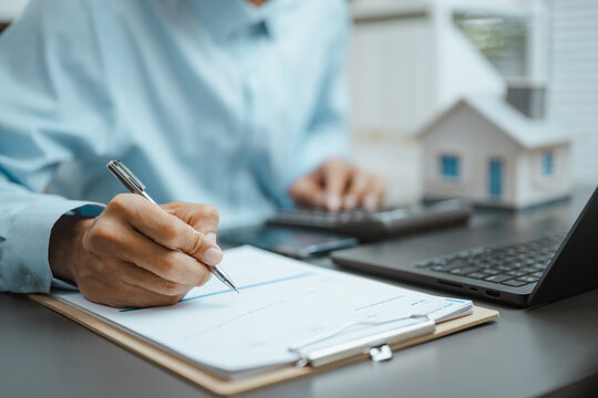 Home model sits beside a person completing forms near a calculator and laptop, illustrating property insurance, mortgage review, and planned investment. Property management, real; property focus.