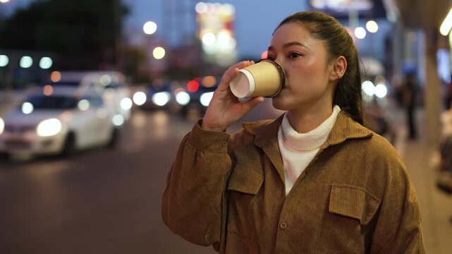 woman holding and drinking a cup of coffee waiting a bus at station in a city street at night