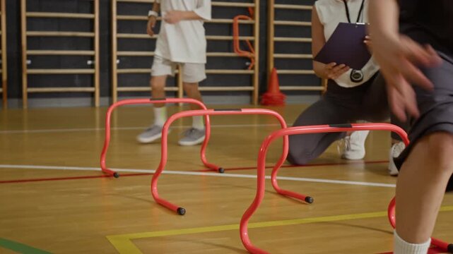 Children jumping over orange hurdles in school gym during physical education class while female coach watching with clipboard. Perfect for youth sports and education marketing