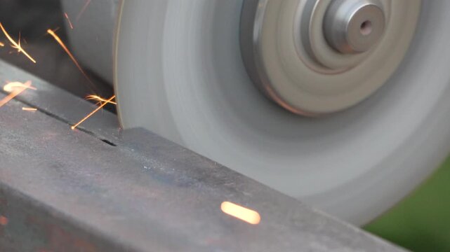 Rotating abrasive blade cuts a dense metal beam while creating glowing sparks outdoors. Macro static shot.