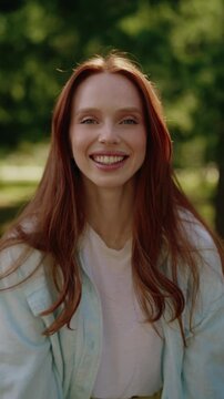Vertical portrait of beautiful young woman with long red hair and radiant smile looking at camera. Cheerful Caucasian model with freckles enjoying sunny day outdoors in green summer park.