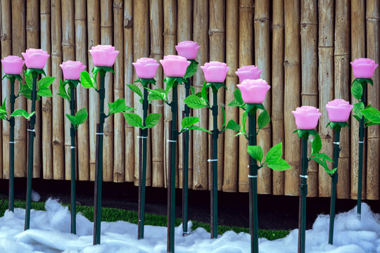 Rows of LED artificial pink rose lights in illuminated garden on artificial snow. LED artificial pink rose lights on dark green stems with fluffy artificial white snow and artificial green grass.