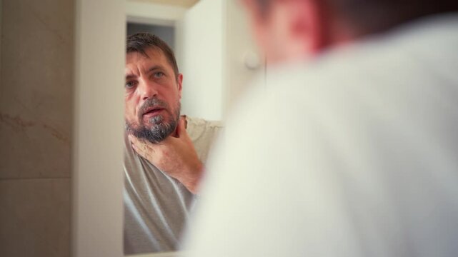 Middle-aged man with graying beard carefully examining his reflection in bathroom mirror and touching his goatee, representing men's grooming, personal care, self-image and morning routine