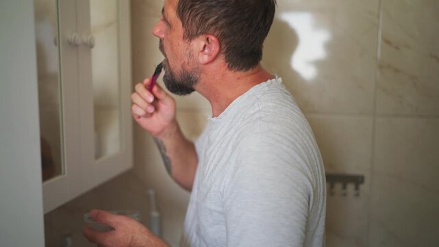 Middle-aged man carefully applying black dye to his graying goatee beard with a pink brush, looking intently at his reflection in the mirror during his home grooming routine