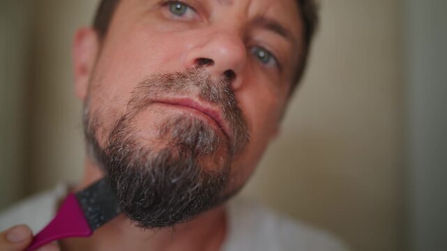 Middle-aged man coloring graying goatee beard with black dye while examining his reflection in mirror, illustrating age-related appearance, grooming and self-care.