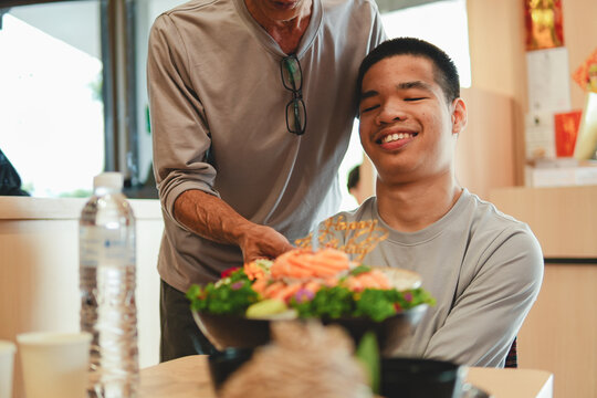 Loving father or caregiver serving a birthday salmon sashimi cake to a happy young man with special needs, enjoying a warm inclusive family celebration together indoors at restaurant or cafe.
