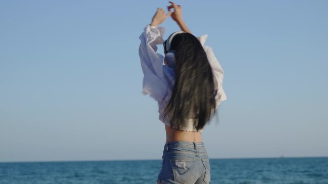 Young woman enjoying music and dancing on sunny ocean shore while wearing modern wireless headphones, white shirt and denim shorts with arms raised against blue sky and sea backdrop.