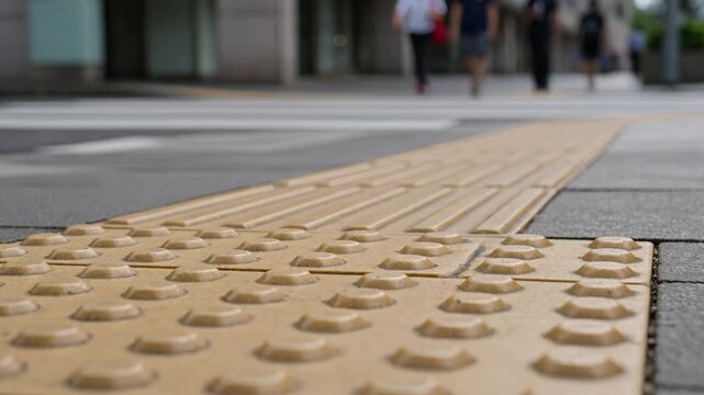 Close-up perspective of yellow tactile paving, also known as tenji blocks, guiding the visually impaired, with people walking on the blurred urban street background