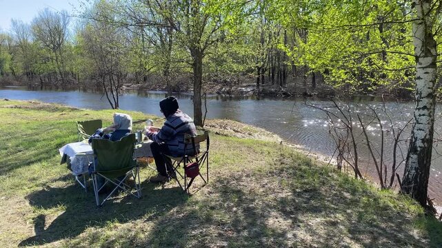 A couple sitting on folding chairs at a picnic table, relaxing on the riverbank and enjoying everyday moments in the fresh air. An atmospheric spot for relaxing in nature