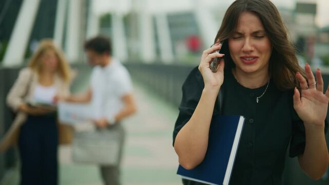 A young businesswoman looks frustrated while talking on her smartphone on a modern bridge. She appears upset during a difficult conversation, holding a notebook in an urban setting.