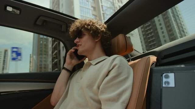 Young adult Caucasian man talking on smartphone while riding in backseat of luxury car, low angle view. Clip is ideal for corporate travel, business commute, and mobile connectivity ads
