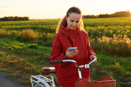 A woman wears a red jacket and stands beside her bicycle on a country road. She is looking at her phone as the sun sets behind the sunflower fields