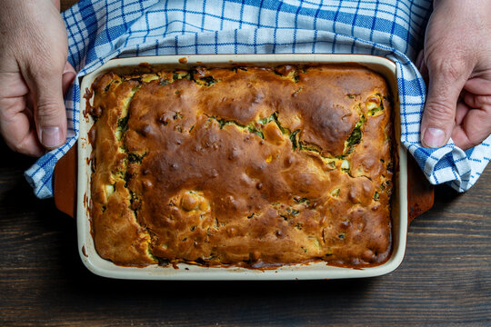 Top view of person holding golden brown savory pie in baking dish with blue checkered towel. Concept of home cooking, family recipe, freshly baked bread or pastry dinner