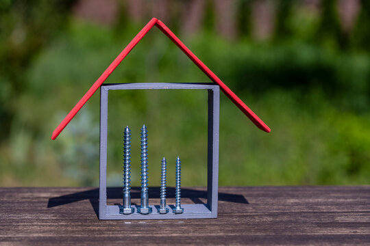 Tiny model house with red roof and silver screws inside on wooden surface against blurred green grass, closeup. Concept of house construction, building materials, mortgage or renovation cost