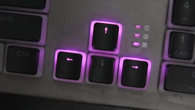 Arrow keys on gaming keyboard with neon purple backlighting close-up. Gamer using his fingers of hand to press key combination while playing computer game in dark room