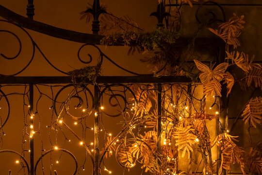 Night festive background with decorated wrought iron fence and shiny garland lights for Halloween and Christmas in autumn style, selective focus. Romantic autumn evening background