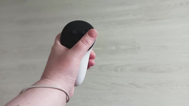 The left hand holding a white virtual reality controller. A virtual reality gamepad with a wrist strap.