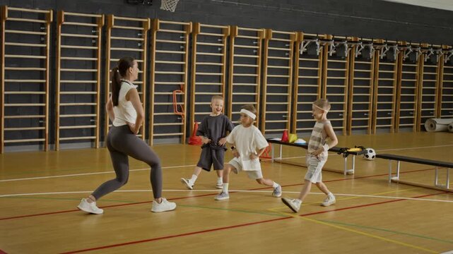 Caucasian female coach demonstrating lunges to three Caucasian kids in school gym. Footage is ideal for promoting youth fitness programs, school sports, or healthy lifestyle campaigns