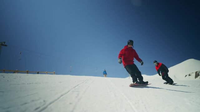 Two snowboarders gracefully carve down a snow-covered mountain slope. They enjoy the crisp air and bright sunshine during their winter sports adventure.