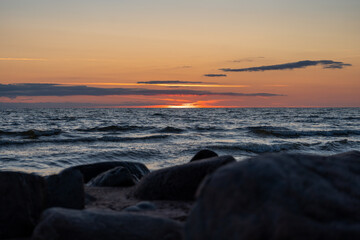 Obraz premium Baltic Sea sunset over gentle waves with dark coastal rocks in the foreground and a dramatic orange evening sky.