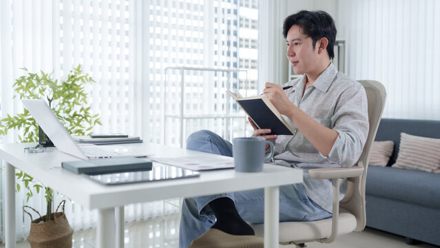 Young Asian man writing in notebook while working from modern home office with city view