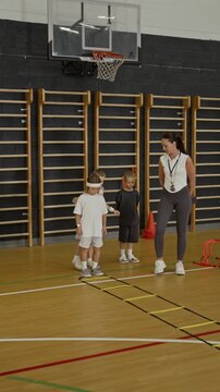 Vertical video of Caucasian female coach teaching group of Caucasian children agility ladder exercises in school gym. Clip is ideal for physical education and youth sports training commercials