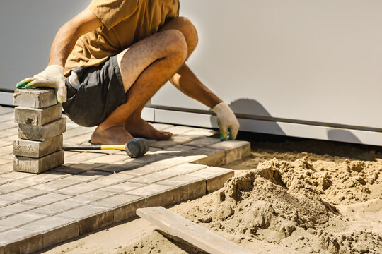 A worker is placing bricks on a prepared surface while kneeling on the ground. Sand and tools are nearby, showing ongoing construction work
