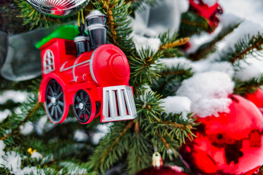 A Christmas tree is decorated with a red train ornament and shiny red baubles. Snow covers the branches, creating a festive atmosphere during New Year