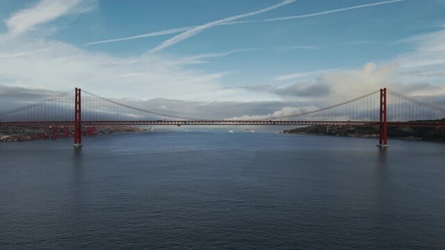 The drone pulls back from 25 de Abril Bridge, showing its red towers against the Tagus River and city.